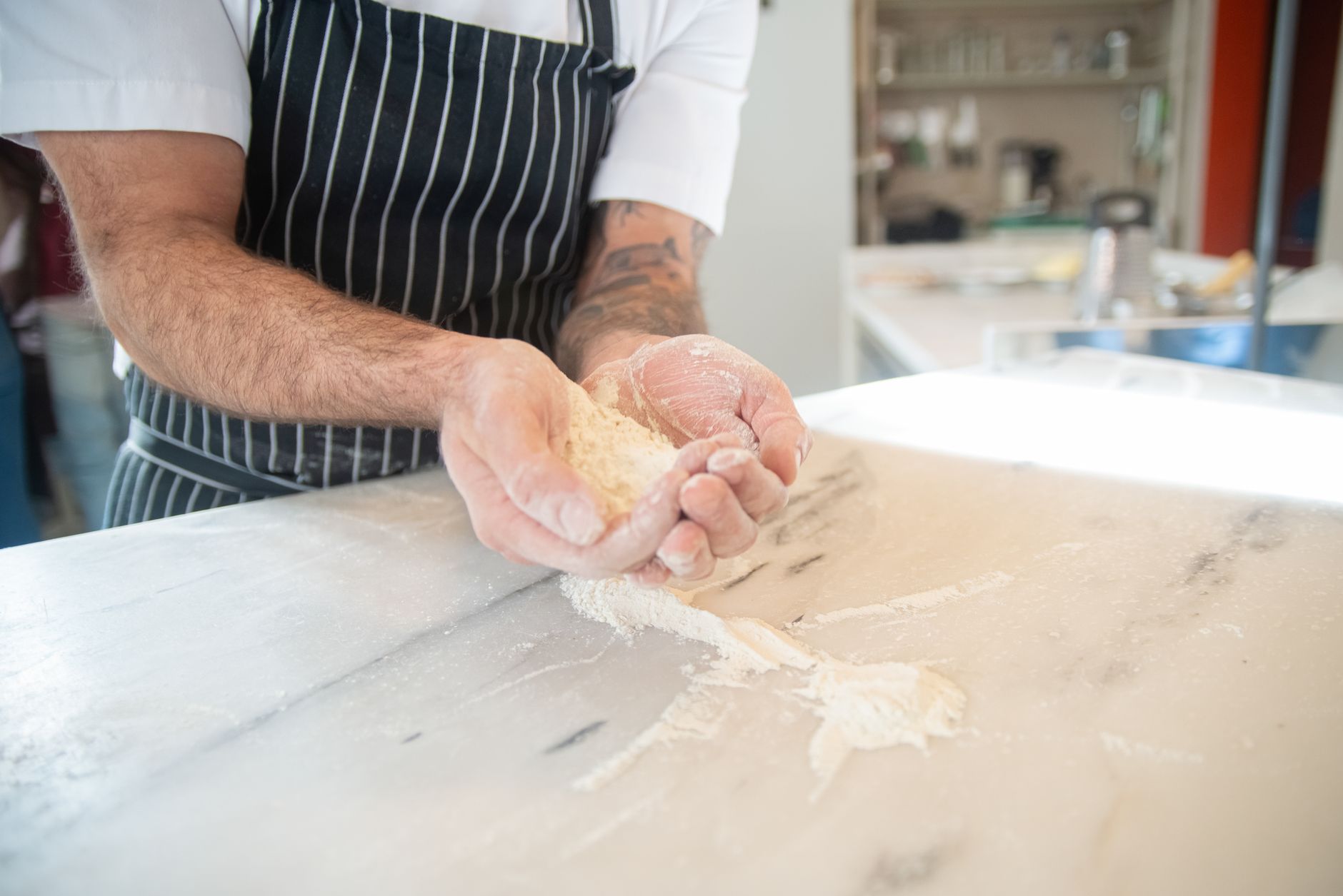 A baker in a striped apron holds flour, preparing dough on a marble countertop.